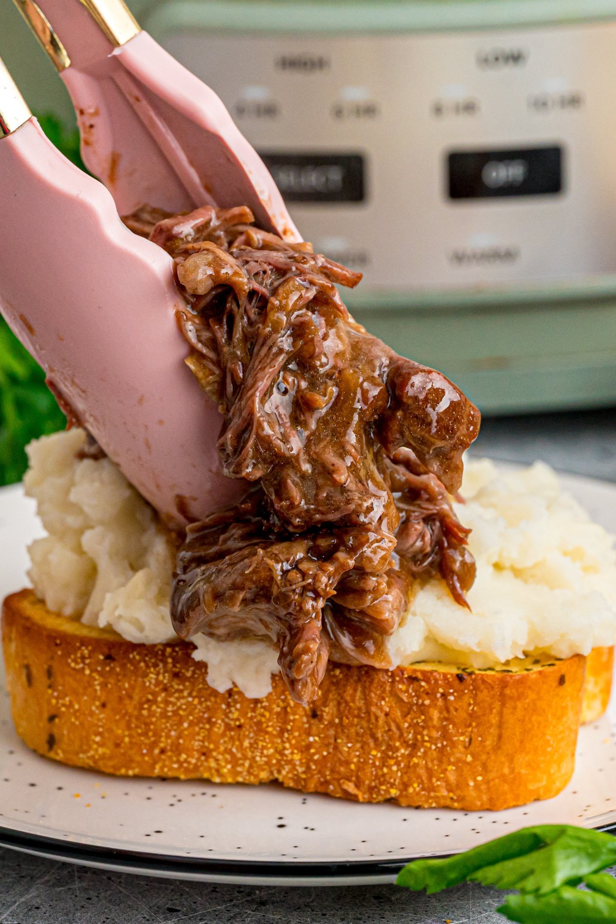 Tongs placing shredded beef on top of mashed potatoes on Texas toast.