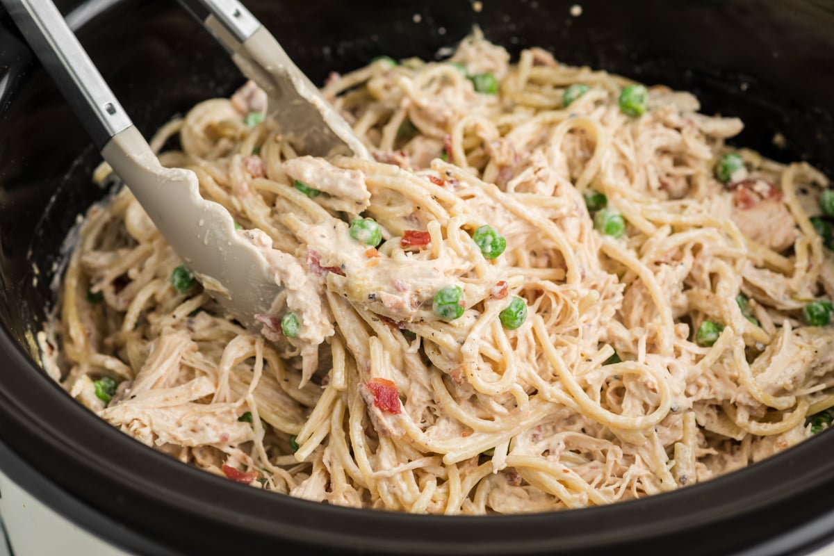 Tongs grabbing some slow cooker chicken carbonara from a crockpot.